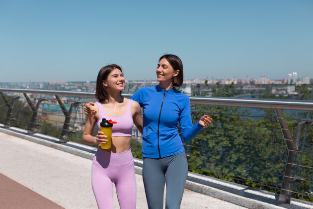 朋友两位穿着运动服的美女在桥上和朋友们愉快而积极地交谈 边走边微笑 享受着健身的早晨 背景上是令人惊叹的城市景色健康运动员运动