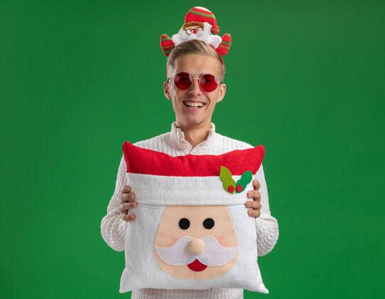 圣诞快乐快乐的年轻帅哥戴着圣诞老人的头带戴着眼镜抱着圣诞老人的枕头看着相机笑着隔离在绿色的背景上空间圣诞老人抱着
