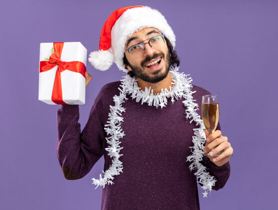 小伙子兴奋的年轻帅哥戴着圣诞帽 脖子上戴着花环 手里拿着一个礼品盒 蓝色背景上隔着一杯香槟花环脖子礼物