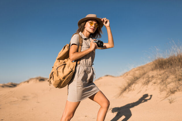 沙滩穿着卡其色衣服的时髦年轻女子在沙漠中漫步 在非洲旅行 戴着帽子和背包 用老式相机拍照游客体验波西米亚