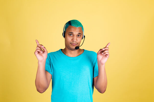 智能休闲黑人非洲男子在休闲黄墙上蓝色头发呼叫中心工作人员快乐的客户支持接线员戴着耳机手指向上服务专业男性