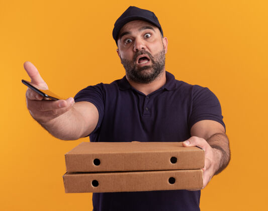 衣服一个身穿制服 戴着帽子 拿着披萨盒的中年送货员吓了一跳 电话就在前面 隔离在黄色的墙上人披萨站着