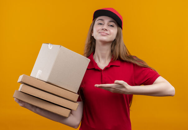 手在孤立的橙色空间里 自信的年轻送货女孩手拿着盒子和包裹 穿着红色制服指着他们橙色盒子红色