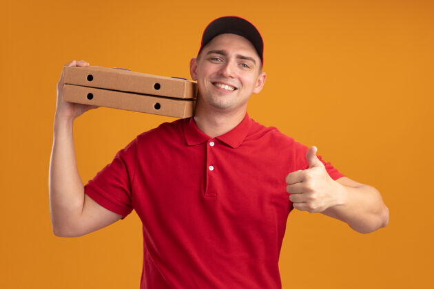 微笑微笑的年轻送货员身穿制服 戴着帽子 肩上扛着披萨盒 在橙色的墙上孤立地竖起大拇指穿着肩膀人