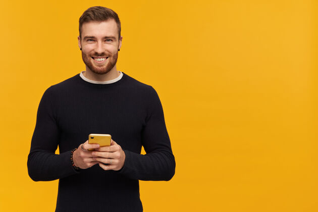 毛衣正面男性的肖像 深色头发 鬃毛有穿孔穿黑色毛衣拿着手机右边的复制空间 隔离在黄色的墙上手机男性服装