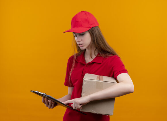 剪贴板困惑的年轻送货女孩穿着红色制服拿着盒子 把手指放在剪贴板上 看着孤立的橙色空间橙色困惑年轻