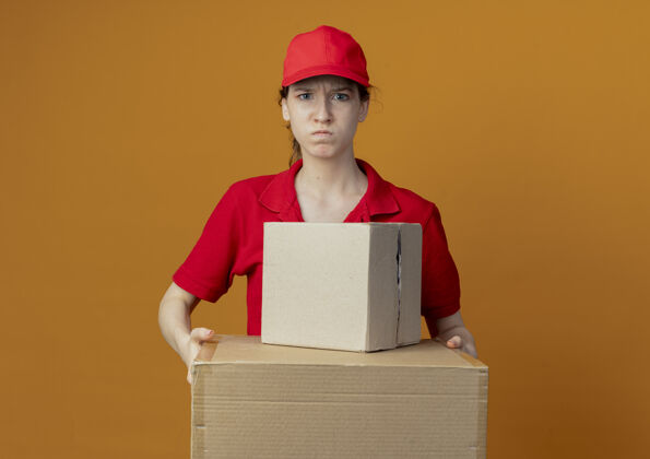 持有愤怒的年轻漂亮的送货女孩 穿着红色制服 戴着帽子 拿着纸箱 看着隔离在橙色背景上的相机 还有复印空间帽子送货年轻