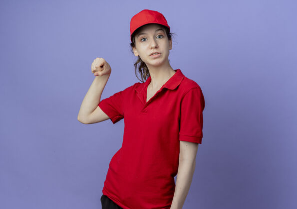 帽子令人印象深刻的年轻漂亮的送货女孩穿着红色制服 戴着帽子假装拿着紫色背景上的复制空间孤立的东西漂亮印象紫色