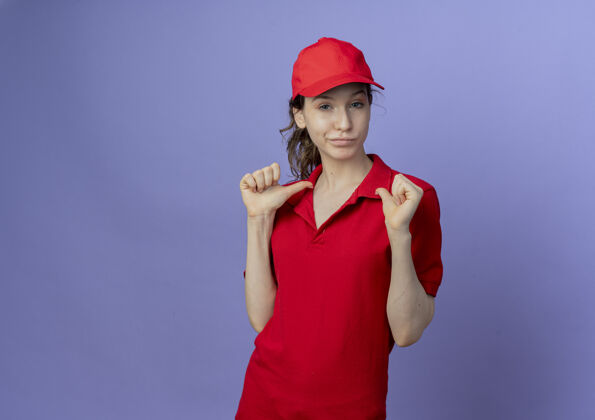 背景自信的年轻漂亮的送货女孩穿着红色制服 戴着帽子 指着自己孤立在紫色背景下的复制空间帽子交付红色