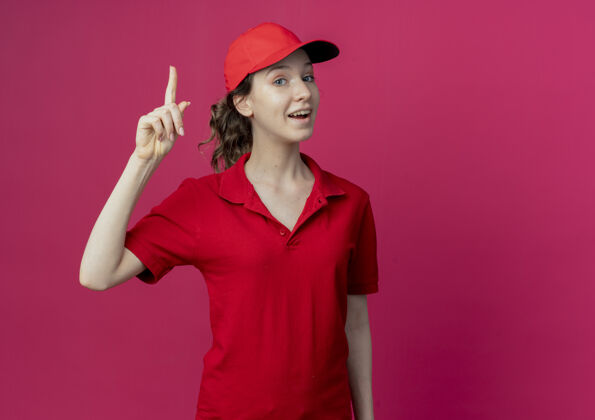 帽子令人印象深刻的年轻漂亮的送货女孩在红色制服和帽子举起手指孤立的深红色背景与复制空间年轻印象交货