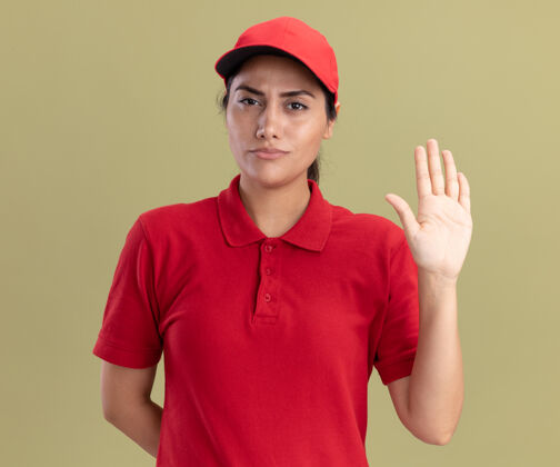 公民严格的年轻送货员穿着制服 戴着帽子 在橄榄绿的墙上显示着停止的手势感情手势脸