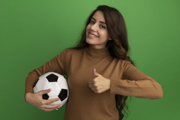 球微笑着看着前面年轻漂亮的女孩拿着球 在绿色的墙上孤立地竖起大拇指姿势人人