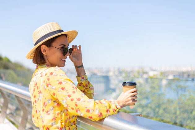 城市穿着黄色夏装 戴着黄色帽子 喝着咖啡 享受阳光的女人的户外肖像 站在桥上 城市的景色令人惊叹旅游者年轻人城镇
