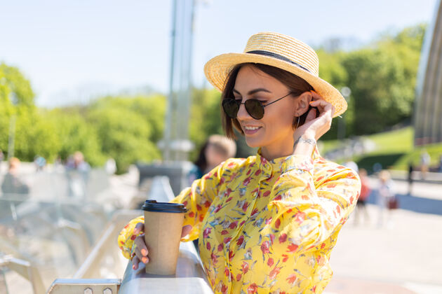 咖啡穿着黄色夏装 戴着黄色帽子 喝着咖啡 享受阳光的女人的户外肖像 站在桥上 城市的景色令人惊叹人女士乐趣