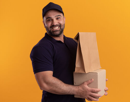 姿势微笑着的中年送货员 穿着制服 戴着帽子 黄色的墙上隔着一个装有纸制食品的盒子人微笑人