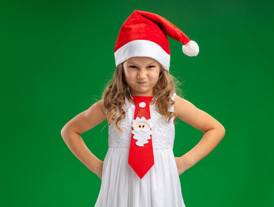 领带愤怒的小女孩戴着圣诞帽打着领带把双手放在臀部隔离在绿色背景上帽子圣诞快乐圣诞节
