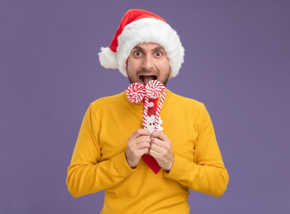 领带印象深刻的年轻白种人戴着圣诞帽 打着领带 拿着圣诞甜甜的手杖在嘴边看着镜头准备吃手杖孤立的紫色背景嘴巴圣诞快乐准备好了