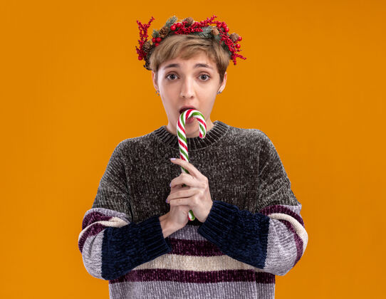 准备好了令人印象深刻的年轻漂亮女孩戴着圣诞花环手持圣诞糖果手杖准备吃它看着隔离在橙色背景上的相机圣诞节橙色头