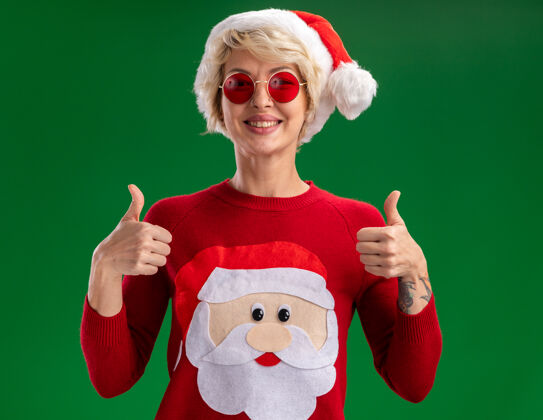 金发快乐的金发女郎戴着圣诞帽 穿着圣诞老人的圣诞毛衣 戴着眼镜 看着相机 在绿色的背景上孤立地竖起大拇指帽子圣诞老人毛衣