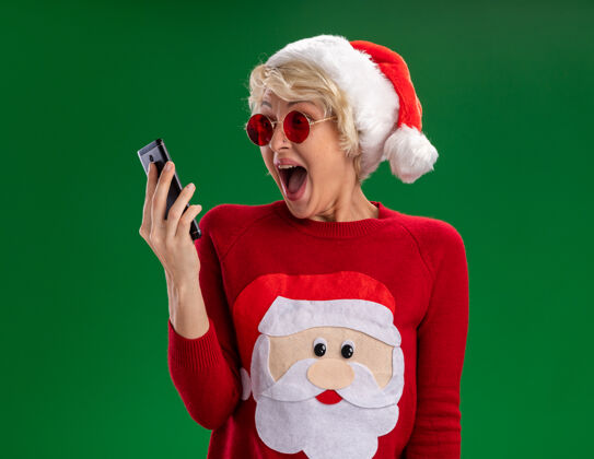 毛衣令人印象深刻的年轻金发女郎戴着圣诞帽和圣诞老人的圣诞毛衣 戴着眼镜 看着绿色背景上孤立的手机眼镜帽子圣诞节