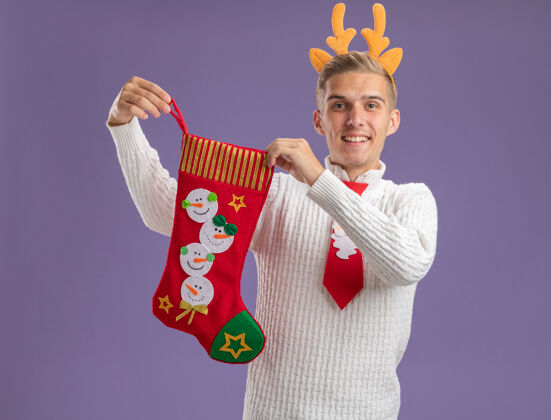 圣诞老人快乐的年轻帅哥戴着驯鹿鹿角头带 打着圣诞老人的领带 手里拿着圣诞长袜 看着隔离在紫色背景上的相机快乐头带年轻人
