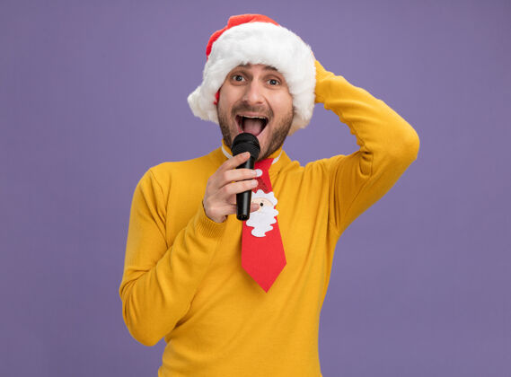 帽子令人印象深刻的年轻白种人戴着圣诞帽打着领带拿着麦克风近嘴看着相机保持手放在头上紫色背景隔离紫色圣诞快乐麦克风