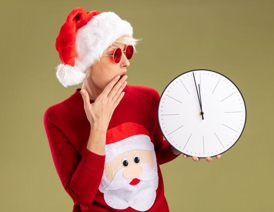 圣诞老人关心的金发女郎戴着圣诞帽 戴着圣诞老人的圣诞毛衣 戴着眼镜 手放在嘴上 看着橄榄绿背景上孤立的时钟圣诞女人眼镜