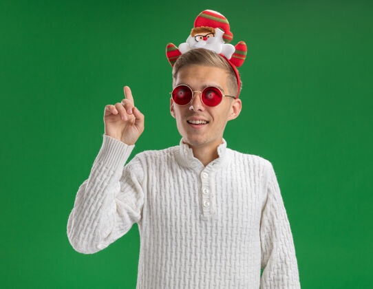圣诞老人给人印象深刻的年轻帅哥戴着圣诞老人的头带 戴着眼镜 一边看着一边 一边指着绿色的背景指向壁板圣诞老人