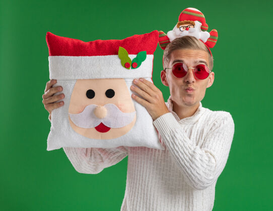 年轻印象深刻的年轻帅哥戴着圣诞老人的头带戴着眼镜抱着圣诞老人的枕头看着镜头做着亲吻的手势隔离在绿色的背景上圣诞老人圣诞老人手势