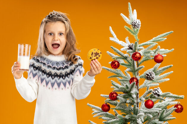 圣诞树快乐的小女孩站在圣诞树旁 戴着头饰 脖子上戴着花环 手里拿着一杯牛奶和饼干 背景是橙色的女孩脖子饼干