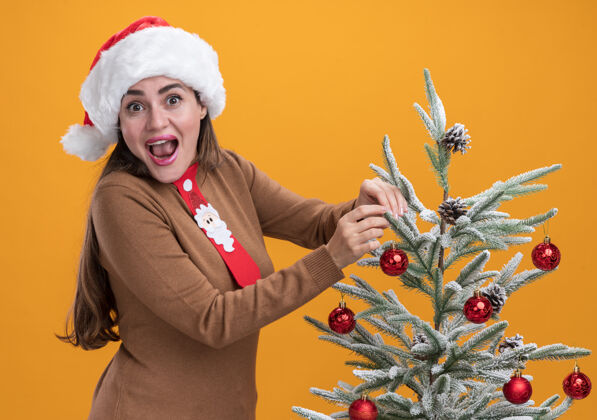 女孩兴奋的年轻漂亮女孩戴着圣诞帽 打着领带 站在橙色背景的圣诞树旁圣诞年轻站起来