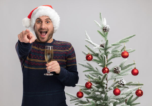 男人令人印象深刻的年轻白种人戴着圣诞帽站在圣诞树旁拿着一杯香槟看着并指着隔离在白色背景上的摄像机香槟帽子举行