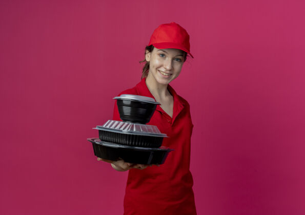 红色面带微笑的年轻漂亮的送货女孩穿着红色制服 戴着帽子 将食品容器伸向摄像机 在深红的背景下与复印空间隔离开来制服帽子送货