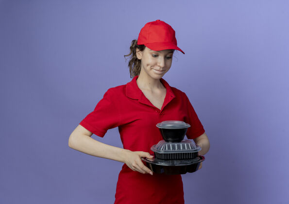 年轻高兴的年轻漂亮的送货女孩穿着红色制服 戴着帽子 看着紫色背景上的食品容器与复制空间隔离紫色容器女孩