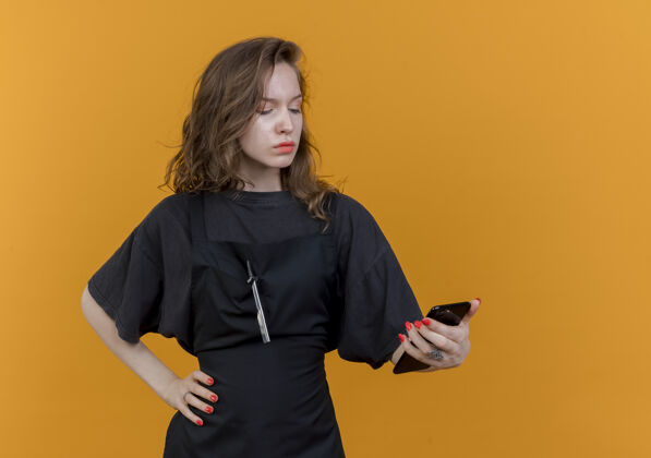手皱眉的年轻斯拉夫女理发师穿着制服拿着手机看着手放在腰上孤立的橙色背景与复制空间腰保持皱眉