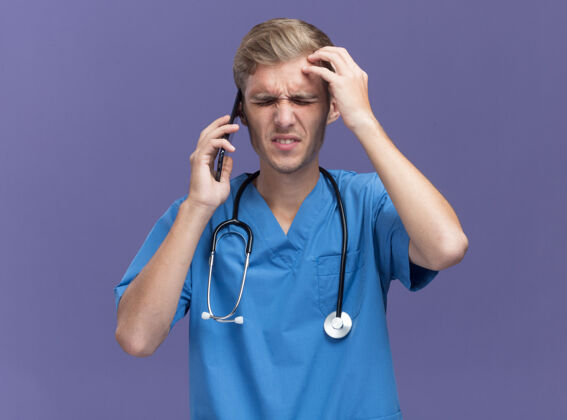 人遗憾地闭上眼睛年轻男医生穿着医生制服用听诊器讲电话把手放在前额隔离在蓝色的墙上后悔眼睛表情