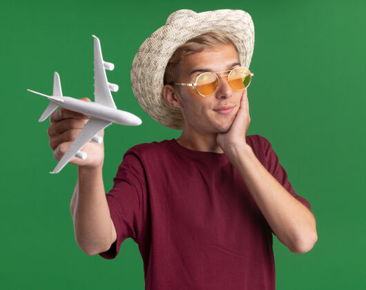 飞机印象深刻的年轻帅哥穿着红衬衫戴着眼镜和帽子拿着玩具飞机把手放在脸颊上隔离在绿色的墙上衣服人帽子