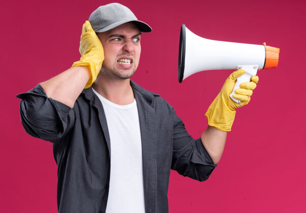 愤怒愤怒的年轻帅气的清洁工 穿着t恤 戴着帽子 戴着手套 看着喇叭 手放在头上 隔离在粉红色的墙上帽子人衣服