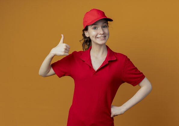 交货微笑着的年轻漂亮的送货女孩 穿着红色制服 戴着帽子 手放在腰上 拇指竖起 孤立地放在橙色背景上 留有复印空间向上女孩腰