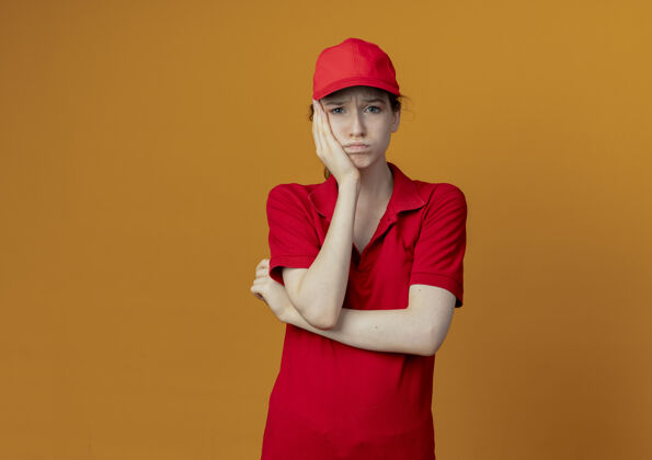 手穿着红色制服 戴着帽子 站在那里 手放在脸上 孤立地放在橙色背景上 留有复印空间 年轻漂亮的送货女孩 未经允许女孩帽子背景