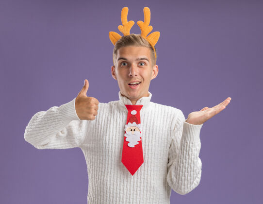 家伙令人印象深刻的年轻帅哥戴着驯鹿鹿角头带和圣诞老人领带看着相机显示空手和拇指向上孤立的紫色背景驯鹿头带印象