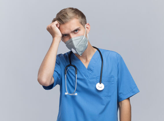公民遗憾的是 年轻的男医生穿着医生制服 戴着听诊器和医用口罩 手放在隔离在白墙上的头上制服姿势面具