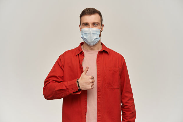 相机自信的年轻胡须男子身穿红衫 脸上戴着防病毒口罩 站在白墙上竖起大拇指年轻医疗面具