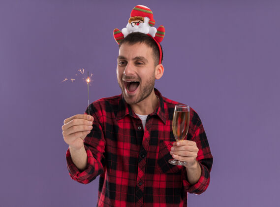 圣诞快乐令人印象深刻的年轻白种人戴着圣诞老人的头带拿着节日火花和一杯香槟看着火花孤立的紫色背景男人圣诞老人印象