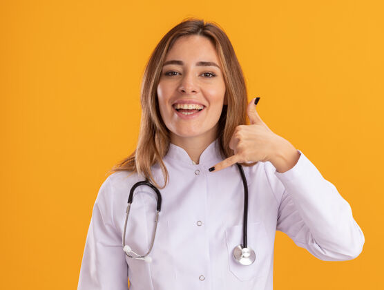 人面带微笑的年轻女医生穿着医用长袍 听诊器显示电话呼叫手势 隔离在黄色墙上微笑电话衣服