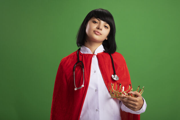 听诊器自信的年轻超级英雄女孩戴着听诊器 穿着医用长袍和披风 戴着王冠穿斗篷长袍