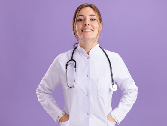 感觉面带微笑的年轻女医生穿着医用长袍 手放在紫色墙上的口袋里脸听诊器微笑