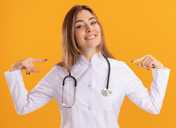年轻人面带微笑的年轻女医生穿着带听诊器的医用长袍 指着隔离在黄色墙上的自己衣服女性她自己