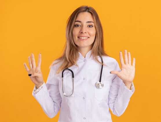 姿势微笑的年轻女医生穿着医用长袍 听诊器显示不同的数字隔离在黄色的墙上医生衣服医学