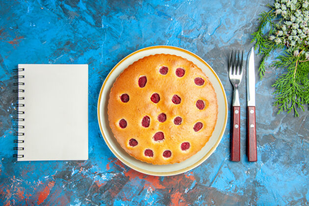 食物树莓蛋糕的俯视图在椭圆形的圆锥形盘子上叉子刀在蓝色的表面上一个笔记本美味刀过滤器
